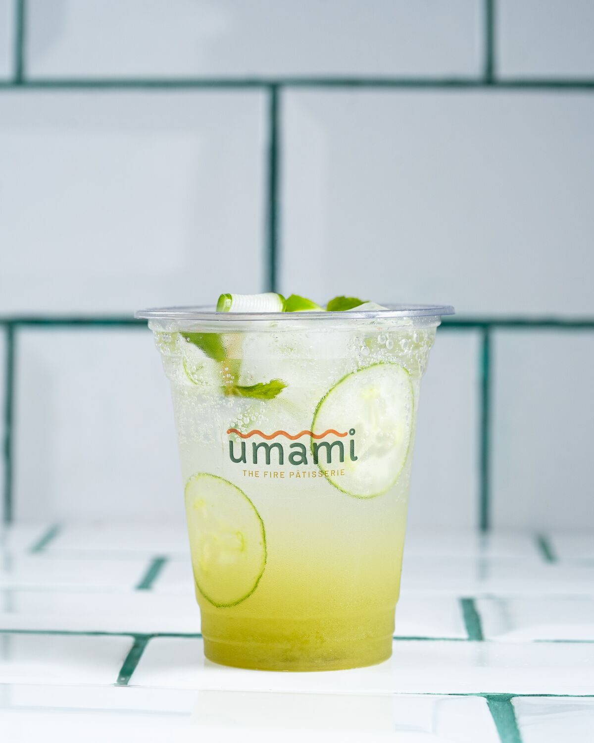 A clear cup of pale green cucumber cooler over ice with fresh cucumber slices and mint leaves.