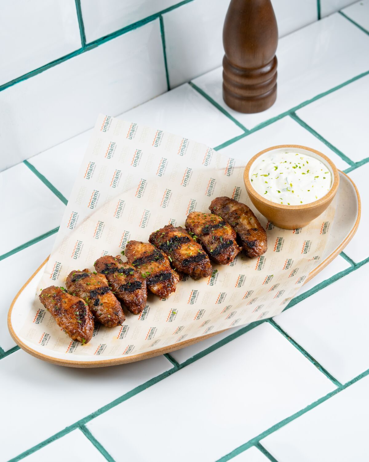 A row of charred fire-grilled chicken wings on a paper lined plate, served with a pot of creamy herb dip.