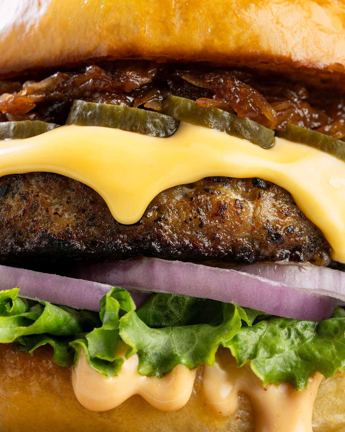 Close up of a charred chicken patty under a thick blanket of melted cheese, layered with pickle slices, caramelised onion, red onion and frilled lettuce, with burger sauce running down the bun.