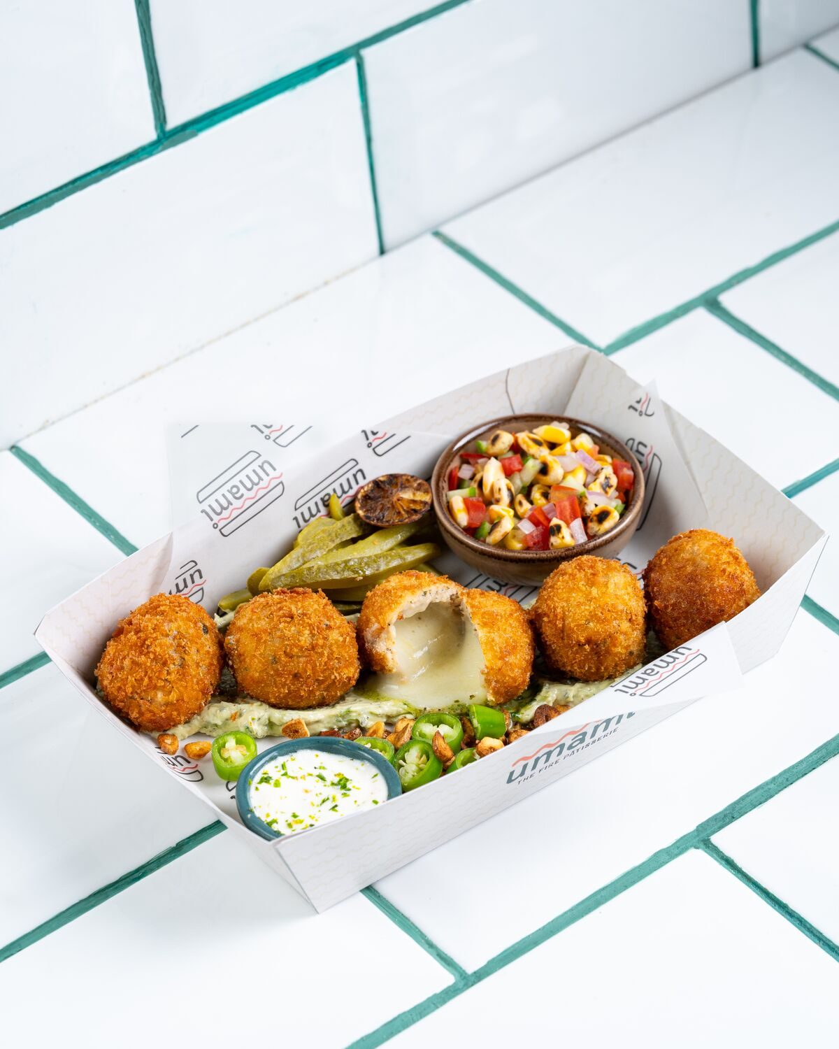 Golden breaded chicken kiev bites in a paper tray on a bed of green herb sauce, one split open to show the molten centre, served with pickles, a corn and pepper salsa and a creamy herb dip.