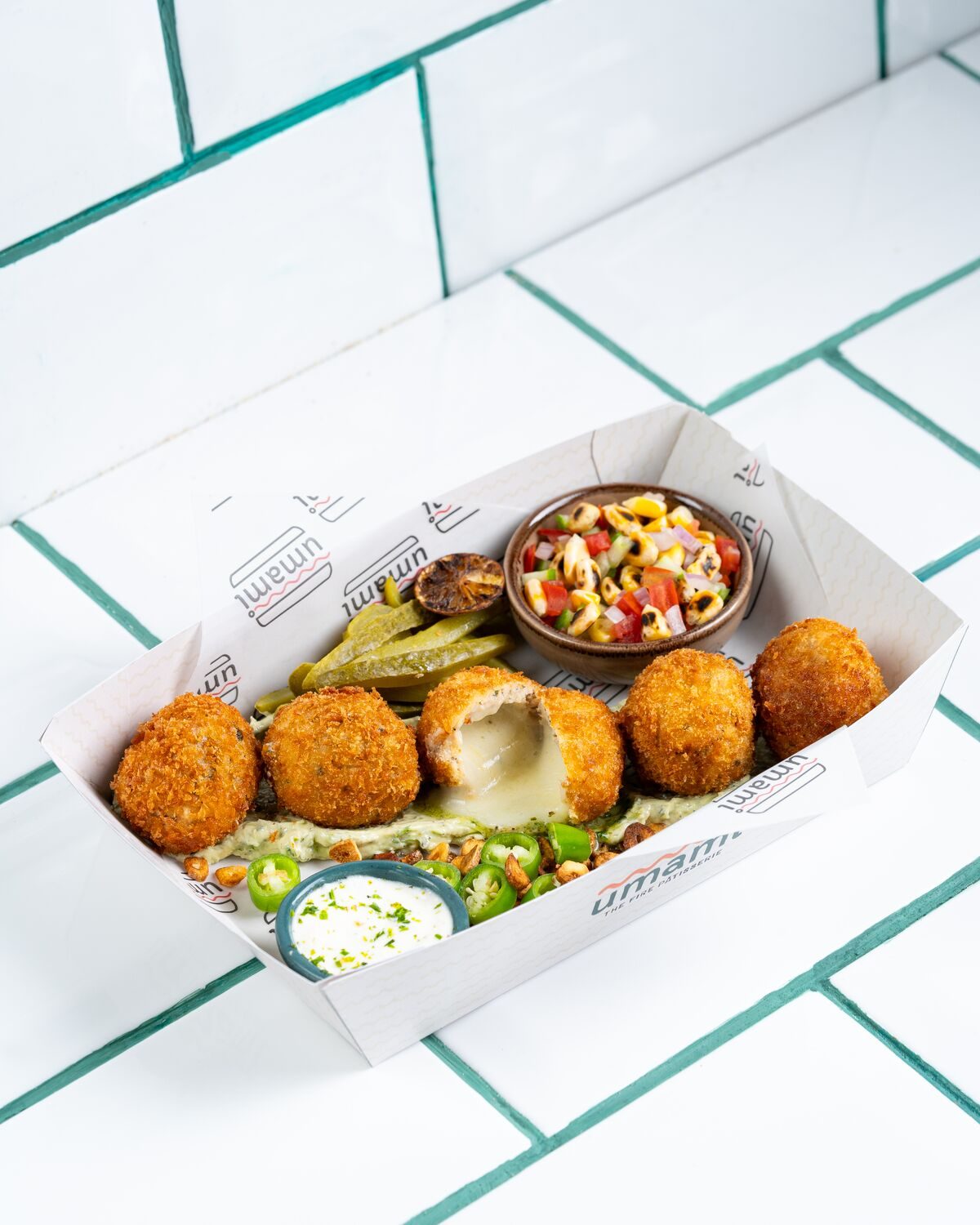 Golden breaded chicken kiev bites in a paper tray, one split open to show the molten garlic butter centre, served with pickles, a herb dip and fresh tomato salsa.