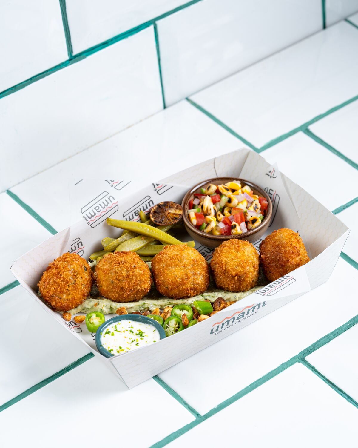Five whole golden breaded chicken kiev bites in a paper tray on a bed of green herb sauce, served with pickles, a corn and pepper salsa and a creamy herb dip.