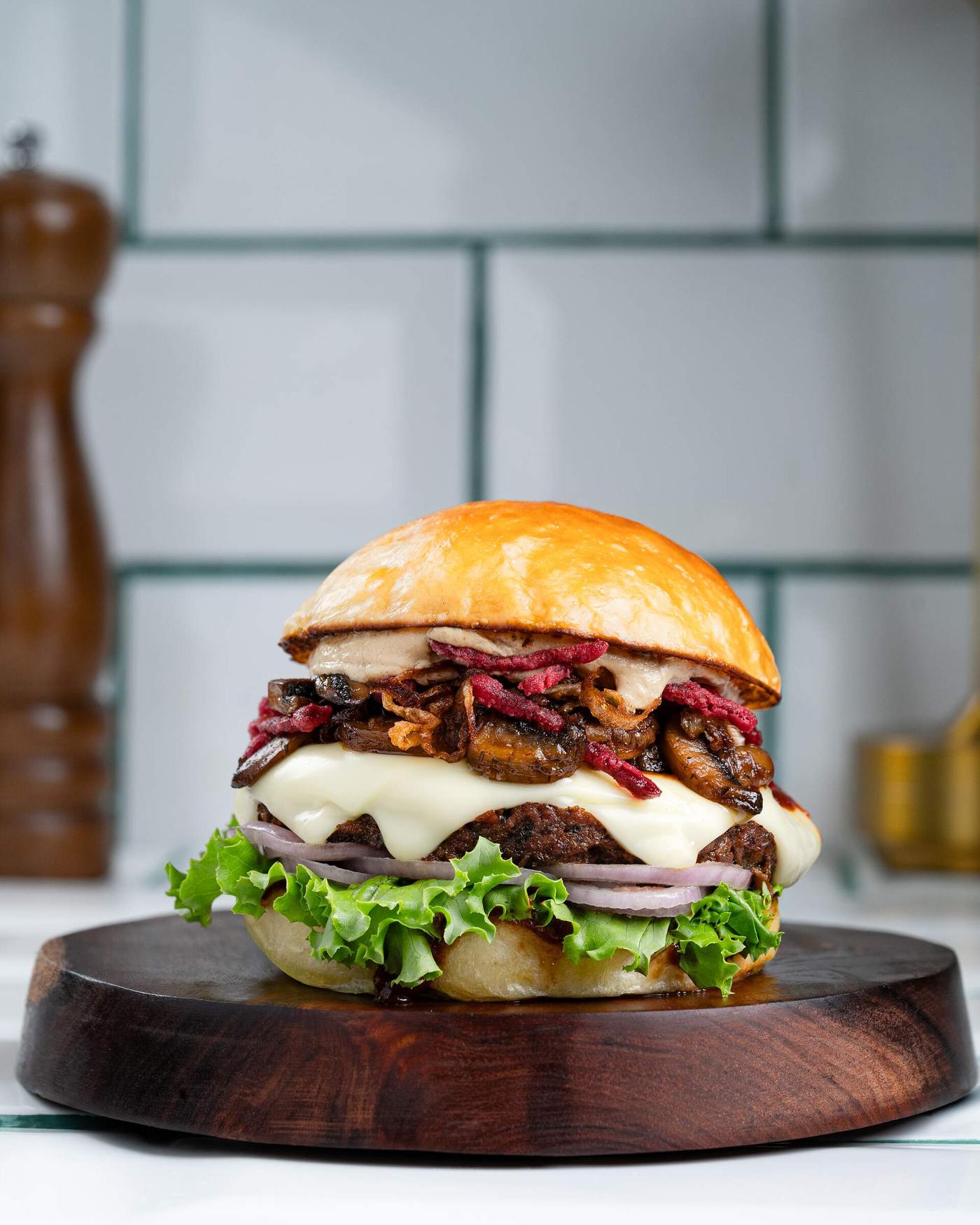 A char-grilled patty covered in melted white cheese and piled with sauteed sliced mushrooms and pickled red onion, with lettuce in a glossy milk bun on a dark wood board.