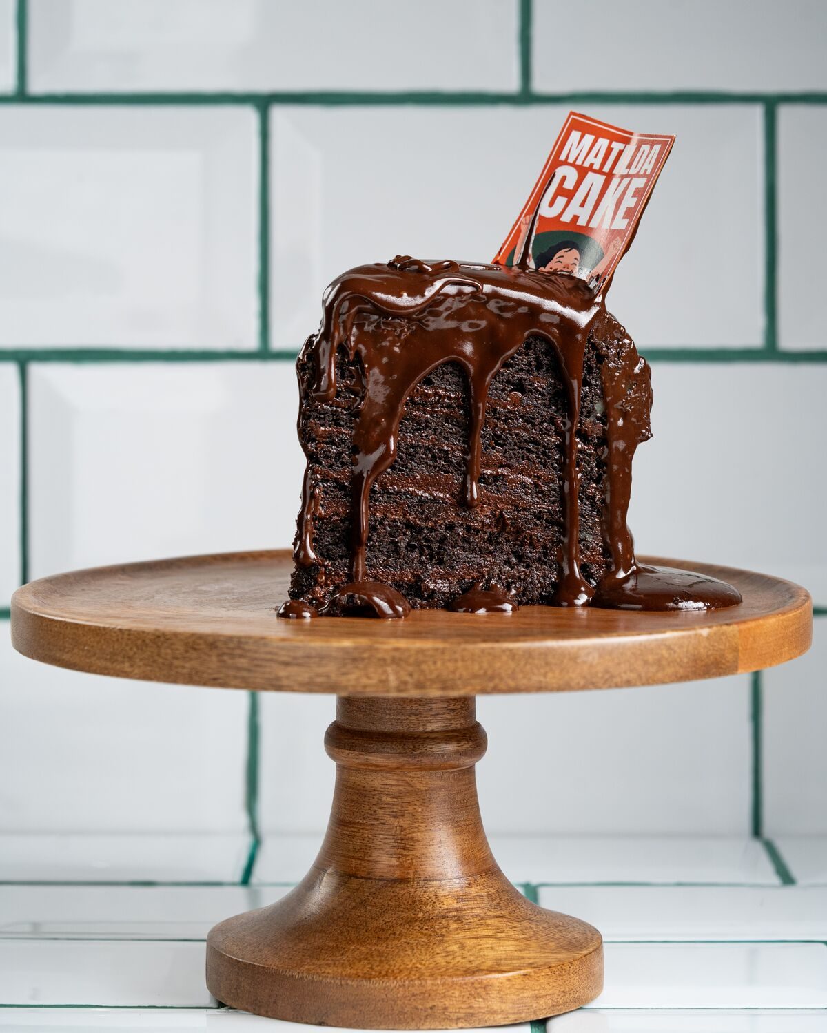 A tall slice of dark chocolate layer cake with thick chocolate ganache pouring down the sides, standing on a wooden cake stand.