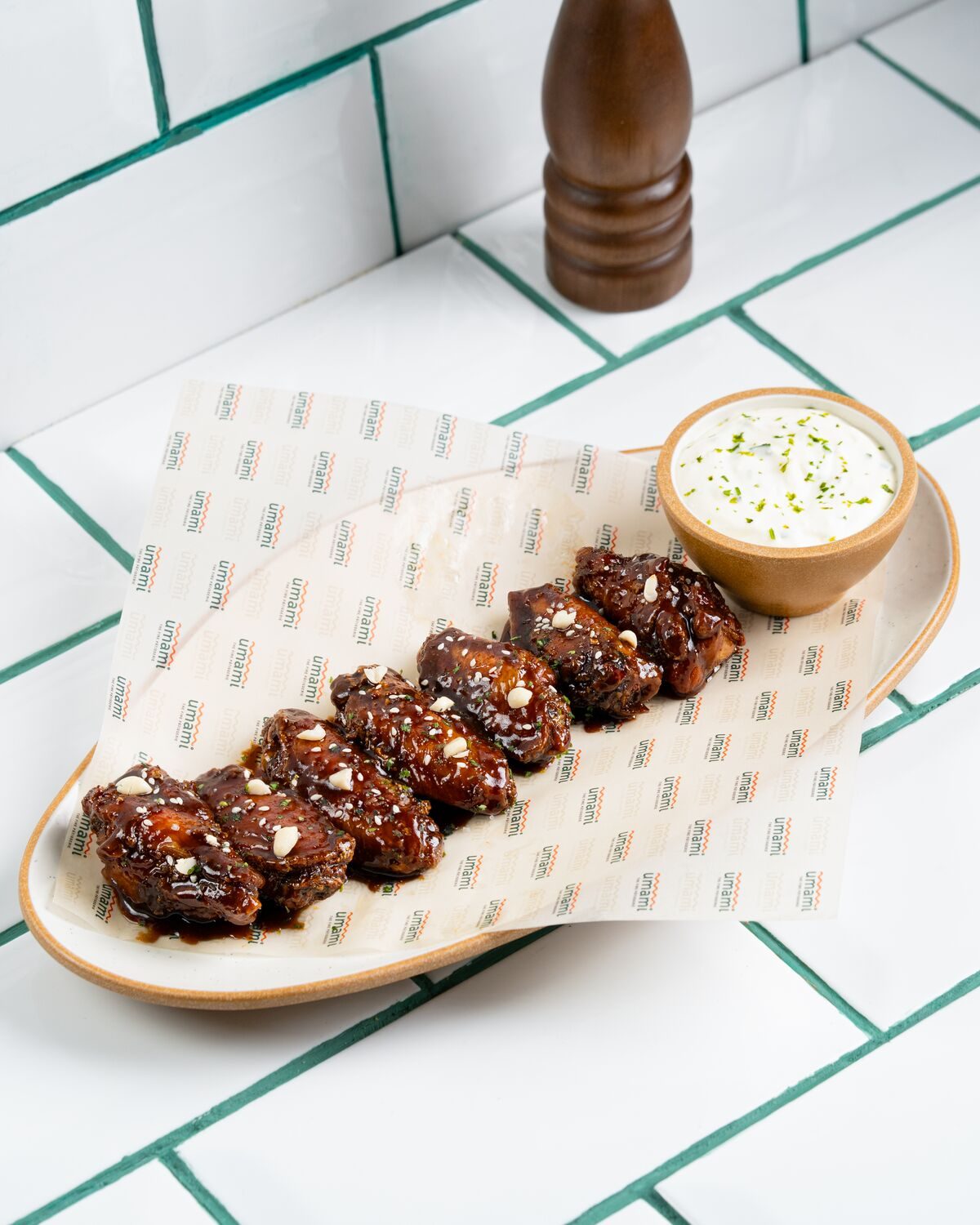 A row of chicken wings in a dark sticky toffee caramel glaze scattered with crushed nuts, served with a pot of creamy herb dip.